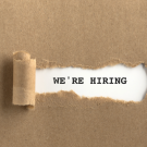 brown paper background, a piece of the paper has been ripped in the center, revealing the words "we're hiring" written on a white surface underneath