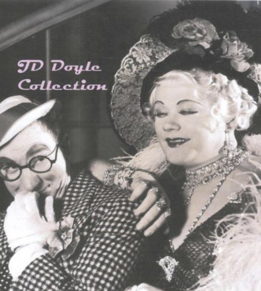Black and white photo from a movie of two men, one dressed in a hat and glasses and the other dressed in drag with a large hat and feather coat.